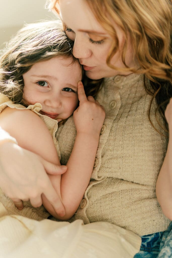 Mother and big sister who is showing a 'number one' with her hand. Snuggled on the couch and mom gives her a kiss on the forehead.