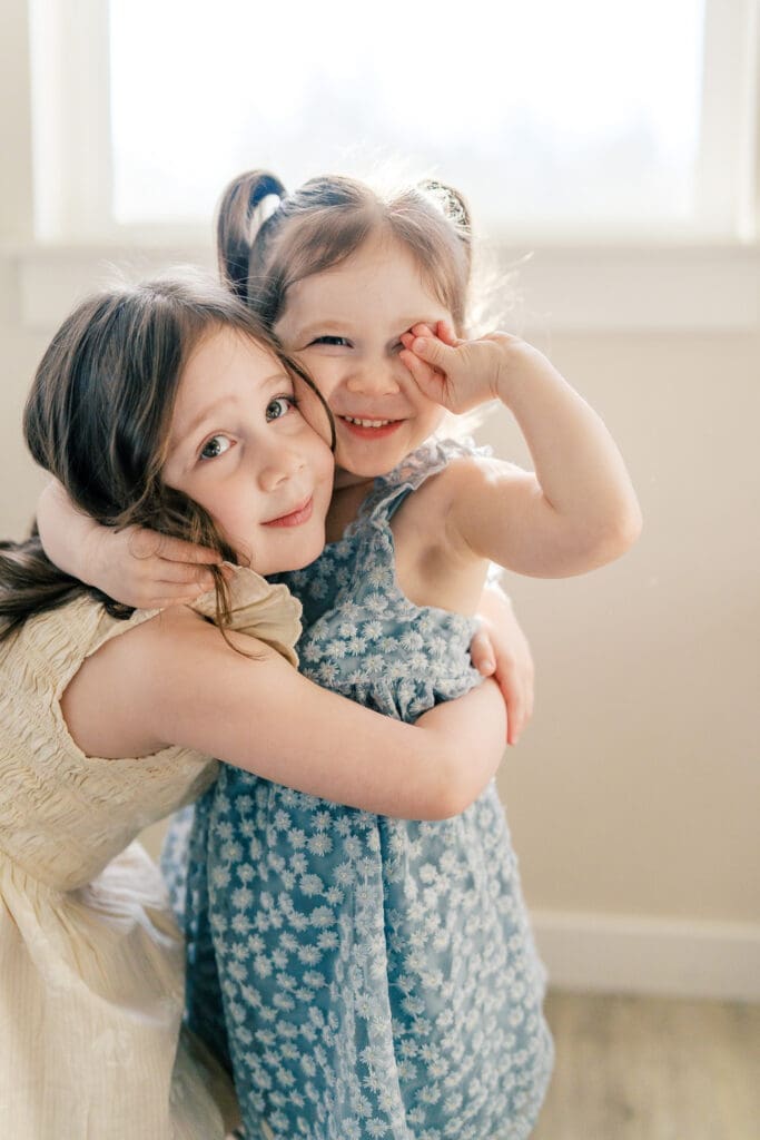 Sisters hugging in a light filled living room, the littlest sister making a silly face