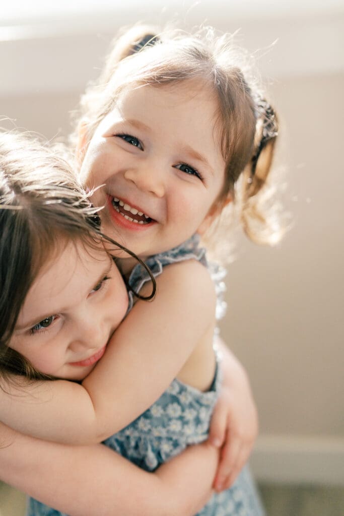Close up of the little sister as she hugs her big sister. Light is pouring into the windows.