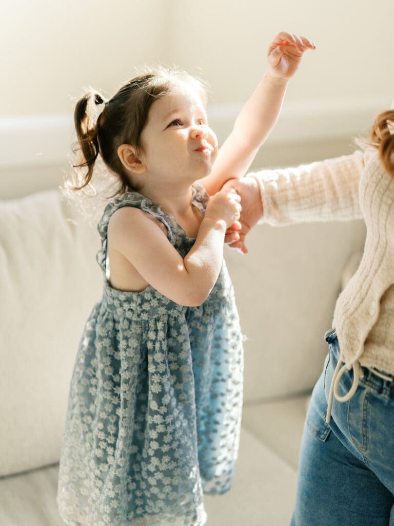 Little sister on the couch reaching for mom while the light pours into the window